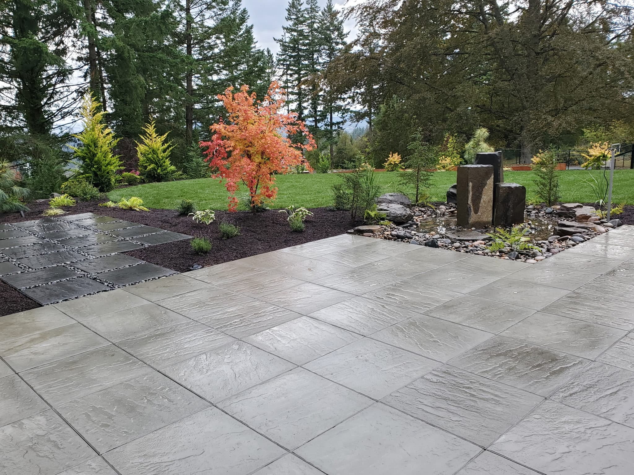 A completed paved patio beside a rock water feature and planted garden beds.